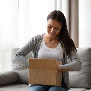 A lady sits on a sofa opening a cardboard box. She is looking into the box and smiling.